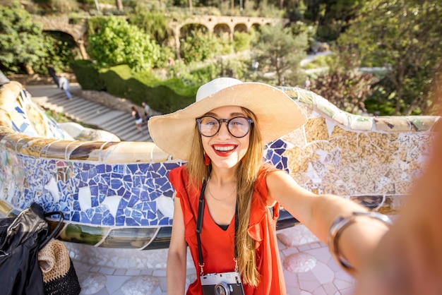 Jeune femme touriste en robe rouge faisant une photo de selfie assise sur le banc décoré de mosaïque dans le célèbre parc Guell à Barcelone