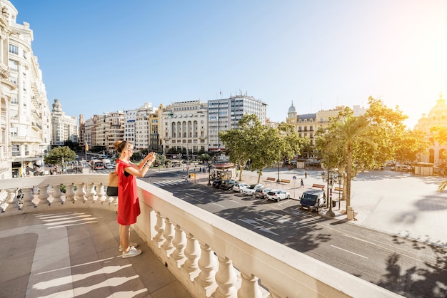 Photo jeune femme touriste en robe rouge photographiant la place ayuntamiento depuis la terrasse de l'hôtel de ville de valence pendant la lumière du matin en espagne