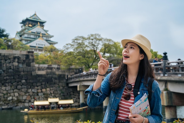 jeune femme touriste tenant une carte papier et trouver la destination. voyageur excité pointant le doigt vers le ciel bleu. rivière d'eau verte propre avec un bateau en bois plein de gens naviguant sur le fond.