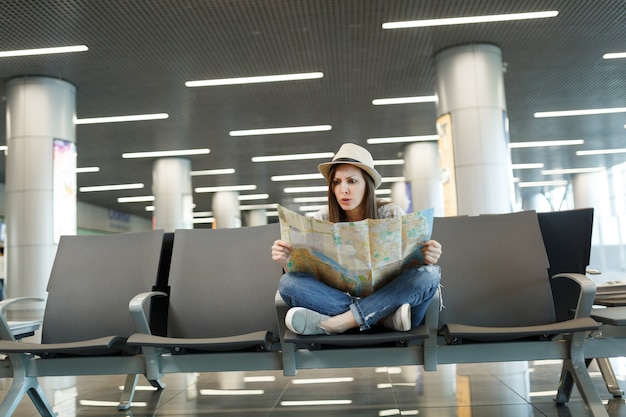 Jeune femme touristique effrayée aux jambes croisées tenant une carte papier, cherchant un itinéraire en attente dans le hall de l'aéroport international