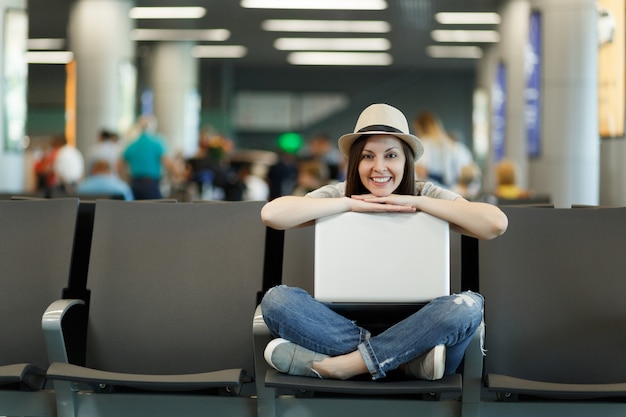 Jeune femme touristique joyeuse voyageuse assise avec un ordinateur portable avec les jambes croisées en attendant dans le hall de l'aéroport international