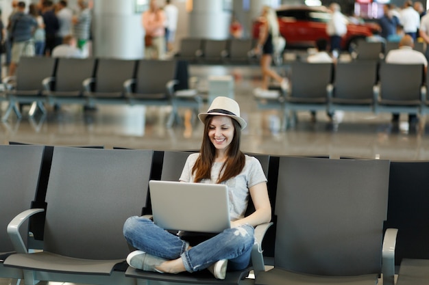 Jeune femme touristique souriante au chapeau assis avec les jambes croisées travaillant sur un ordinateur portable attendre dans le hall de l'aéroport international