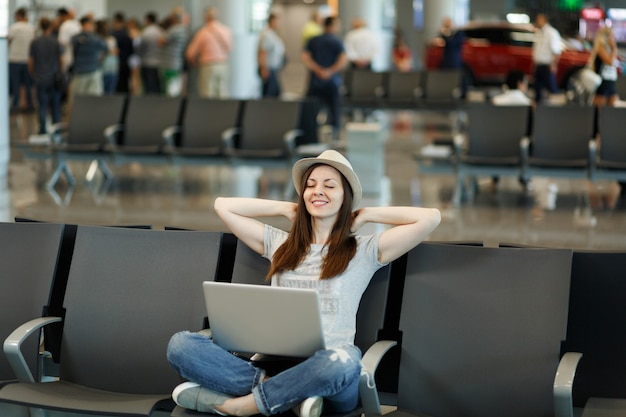 Jeune femme touristique voyageur détendue au chapeau assis avec les jambes croisées travaillant sur un ordinateur portable attendre dans le hall de l'aéroport international