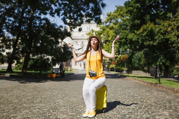 Jeune femme touristique de voyageur insatisfait en vêtements jaunes assis sur une valise tenant une carte de la ville écartant les mains en plein air. Fille voyageant à l'étranger pour voyager en week-end. Mode de vie de voyage touristique.