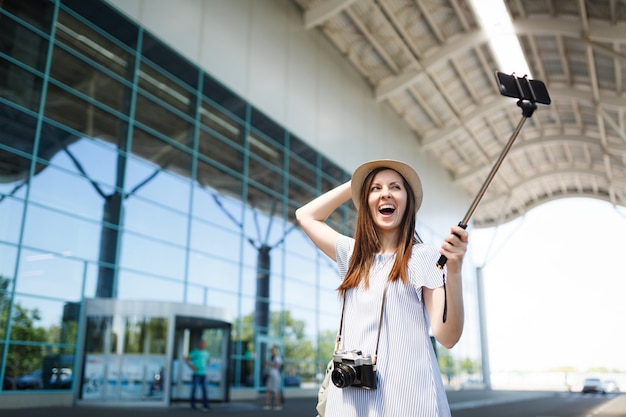 Jeune femme touristique voyageuse ravie avec un appareil photo vintage rétro faisant un selfie sur un téléphone portable avec un bâton égoïste monopode à l'aéroport