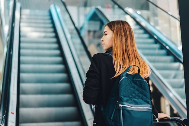 Jeune femme touristique yong rousse avec un sac à dos approchant d'un escalator dans le terminal de l'aéroport international