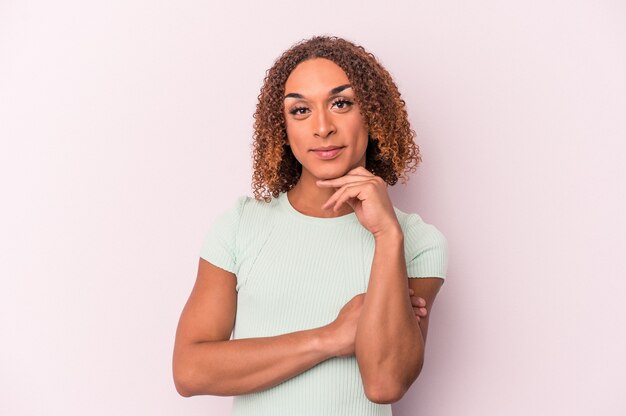 Jeune femme transsexuelle latine isolée sur fond rose souriante heureuse et confiante, touchant le menton avec la main.