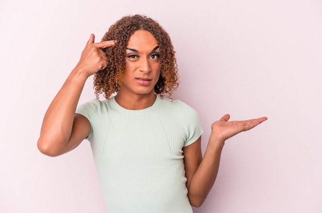 Jeune femme transsexuelle latine isolée sur fond rose tenant et montrant un produit à portée de main.