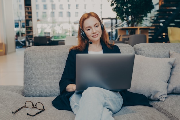 Jeune femme travaillant comme agent de centre d'appels en ligne, communique avec les clients à l'aide d'écouteurs sans fil et d'un ordinateur portable, travaillant à distance sur une hotline d'assistance tout en étant assise sur un canapé dans un café