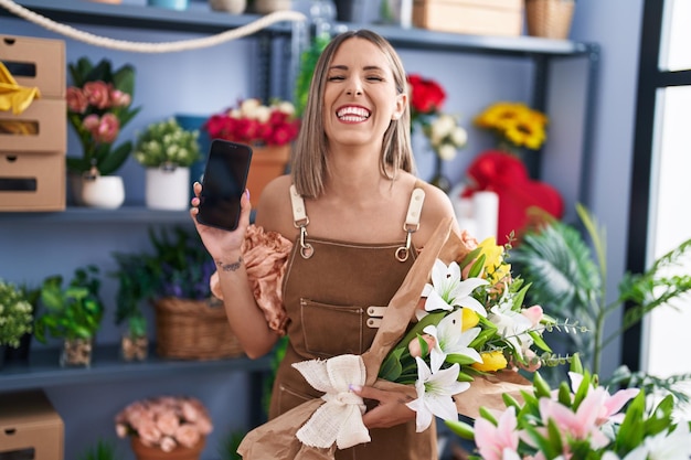 Jeune femme travaillant dans un fleuriste montrant un écran de smartphone souriant et riant fort à haute voix parce qu'une drôle de blague folle.