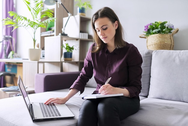 Jeune femme travaillant à distance à la maison, femme d'affaires indépendante assise sur un canapé avec un ordinateur portable, prenant des notes dans un cahier d'affaires