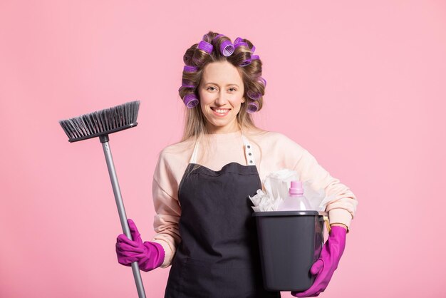 Une jeune femme travaillant à domicile arrange les cheveux avec des rouleaux portant un tablier tenant un balai pour balayer le sol et un seau de vadrouille contenant du liquide