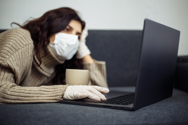 Jeune femme travaillant à domicile pendant la quarantaine en raison d'une pandémie de coronavirus. Belle fille reste à la maison avec un masque médical et des gants et en tapant sur un ordinateur portable. Concept mondial de l'épidémie de Covid-19.