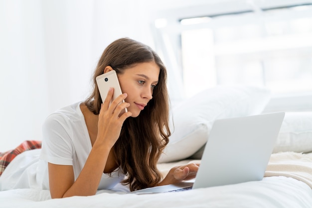 Jeune femme travaillant à la maison portant sur le lit en pyjama, parlant sur mobile. Femme dans la chambre