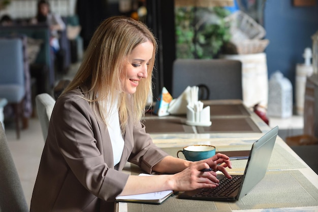 Une jeune femme travaille en ligne sur un ordinateur portable dans un café au déjeuner à côté d'un cahier une tasse de café