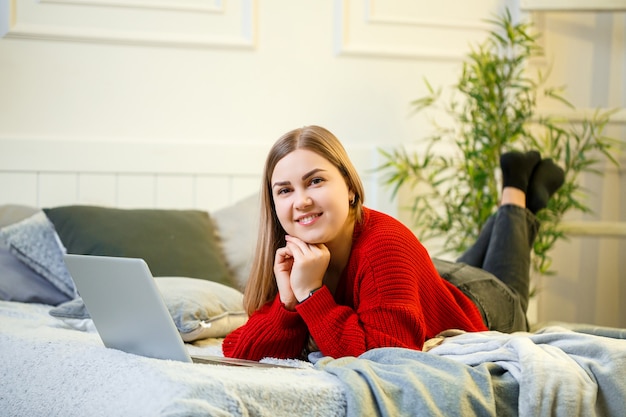 Jeune femme travaille à un ordinateur, assise sur un lit, travaillant à distance. Une fille aux cheveux longs vêtue d'un pull rouge et d'un jean travaille à la maison.