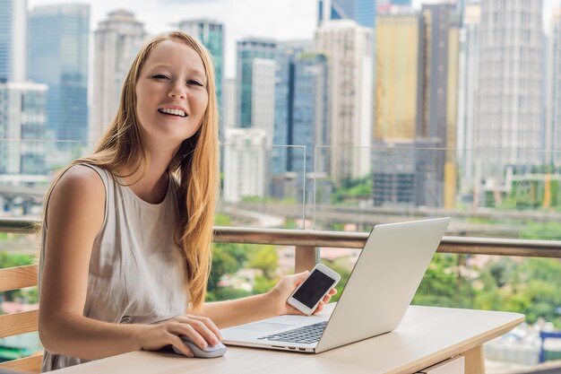 Une jeune femme travaille sur un ordinateur portable sur son balcon donnant sur le travail à distance d'un pigiste de gratte-ciel