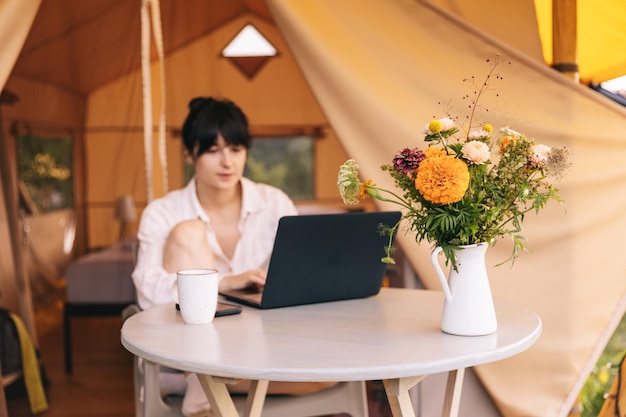 Une jeune femme travaille sur un ordinateur portable tout en étant assise détendue sur une chaise près du feu de camp voyageant avec une tente Concept de travail à distance et d'évasion dans la nature