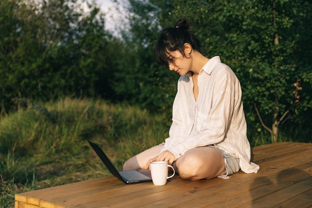 Une jeune femme travaille sur un ordinateur portable tout en étant assise détendue sur une chaise près du feu de camp voyageant avec une tente Concept de travail à distance et d'évasion dans la nature