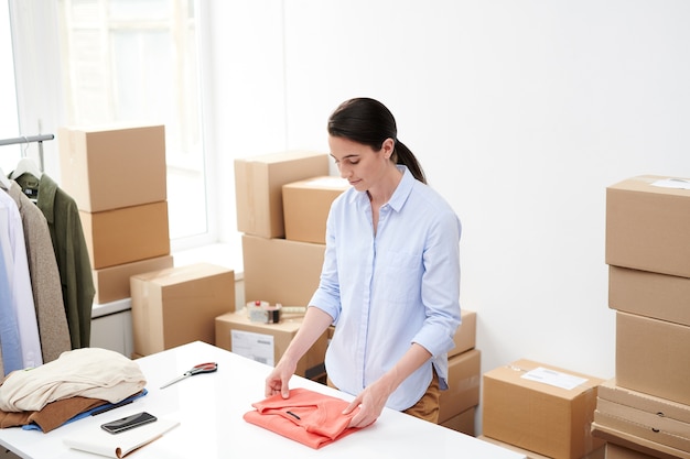 Jeune femme travailleur de la boutique en ligne pliant pull rouge pour l'emballage tout en se tenant un bureau au bureau