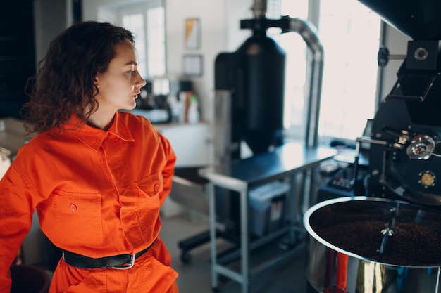 Photo jeune femme travailleuse en costume orange en atelier avec torréfacteur de café pendant le processus de torréfaction du café