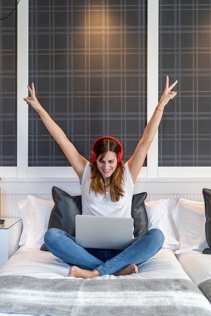 Jeune femme très heureuse avec ses bras assis sur son lit en écoutant de la musique et en travaillant sur son ordinateur portable