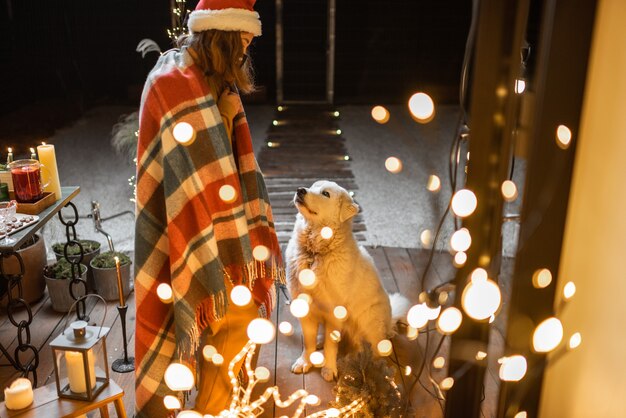 Jeune femme triste au masque facial célébrant avec un chien les vacances du Nouvel An sur une terrasse à la maison. Concept de quarantaine et d'auto-isolement pendant l'épidémie en vacances