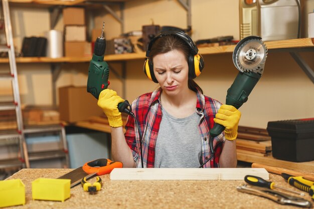 Jeune femme triste bouleversée en chemise à carreaux, T-shirt gris, écouteurs insonorisés, gants jaunes travaillant dans un atelier de menuiserie sur une table en bois avec différents outils, scie électrique et perceuse électrique.