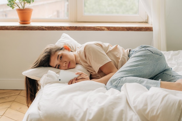Une jeune femme triste regardant son smartphone pendant la journée alors qu'elle est allongée sur le lit dépendance à Internet