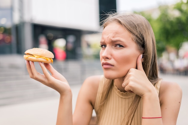 Jeune femme triste tenant un hamburger assis à l'extérieur