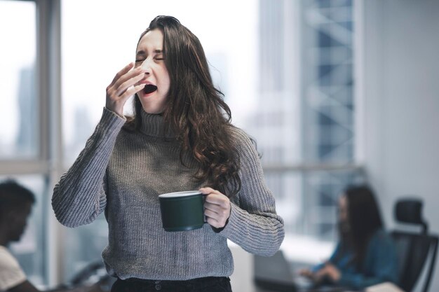 Jeune femme turque désordonnée et fatiguée dans un bureau moderne avec ses collègues Elle bâille et tient une tasse de café