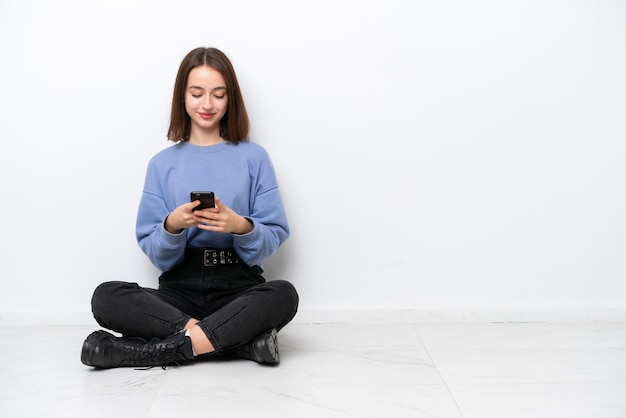 Jeune femme ukrainienne assise sur le sol isolé sur fond blanc envoyant un message avec le mobile