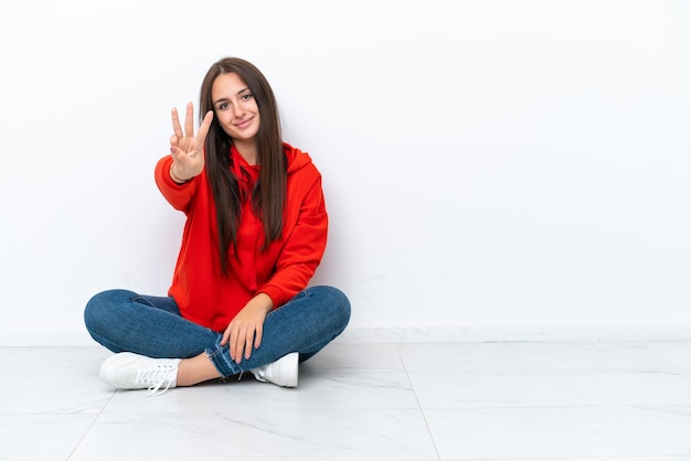 Jeune femme ukrainienne assise sur le sol isolé sur fond blanc heureux et comptant trois avec les doigts