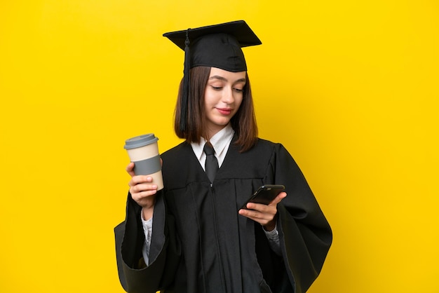 Jeune femme ukrainienne diplômée universitaire isolée sur fond jaune tenant du café à emporter et un mobile