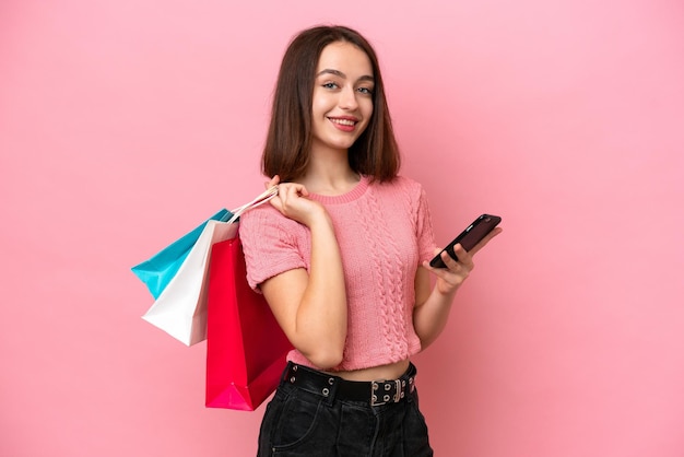 Jeune femme ukrainienne isolée sur fond rose tenant des sacs à provisions et écrivant un message avec son téléphone portable à un ami