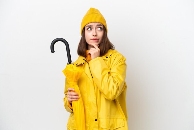 Jeune femme ukrainienne avec manteau imperméable et parapluie isolé sur fond blanc ayant des doutes et pensant