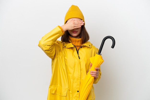 Jeune femme ukrainienne avec manteau imperméable et parapluie isolé sur fond blanc couvrant les yeux par les mains Je ne veux pas voir quelque chose