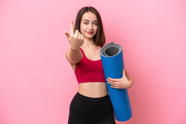 Jeune femme ukrainienne sportive allant à des cours de yoga tout en tenant un tapis isolé sur fond rose faisant un geste à venir