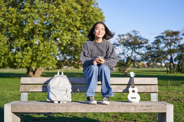 Jeune femme avec ukulele assis sur un banc dans le parc à l'aide d'une application de téléphonie mobile tenant un smartphone et un sourire