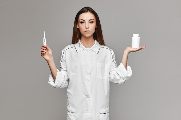 Jeune femme en uniforme médical tenant un pot de pilules dans une main et un thermomètre médical dans une autre et posant sur le fond gris. Concept de soins de santé et de médecine