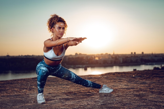 Jeune femme urbaine faisant des exercices d'étirement au bord de la rivière au coucher du soleil.