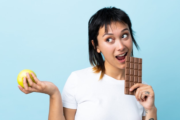 Jeune femme uruguayenne sur fond bleu isolé prenant une tablette de chocolat dans une main et une pomme dans l'autre