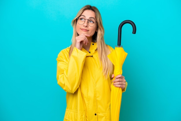 Jeune femme uruguayenne avec manteau imperméable et parapluie isolé sur fond bleu et regardant vers le haut