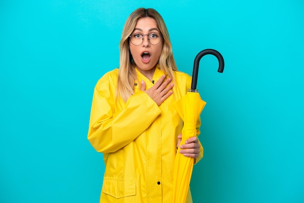 Jeune femme uruguayenne avec manteau imperméable et parapluie isolé sur fond bleu surpris et choqué tout en regardant à droite