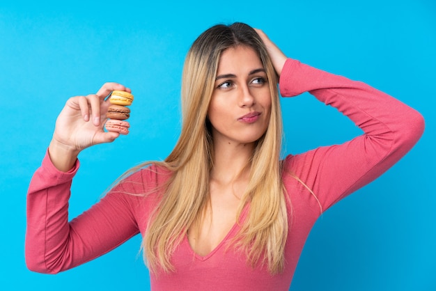 Jeune femme uruguayenne sur mur bleu isolé tenant des macarons français colorés avec une expression confuse