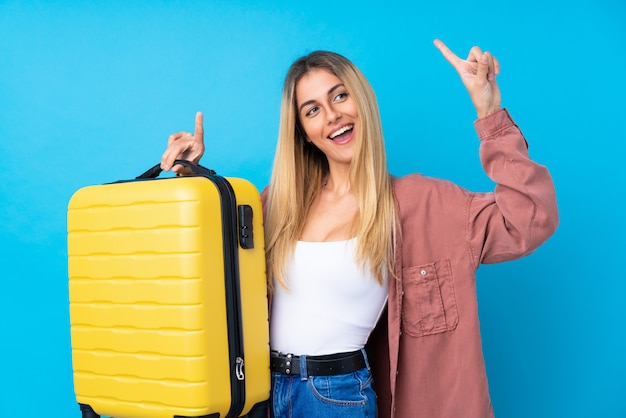 Jeune femme uruguayenne sur mur isolé mur bleu en vacances avec valise de voyage et pointant vers le haut