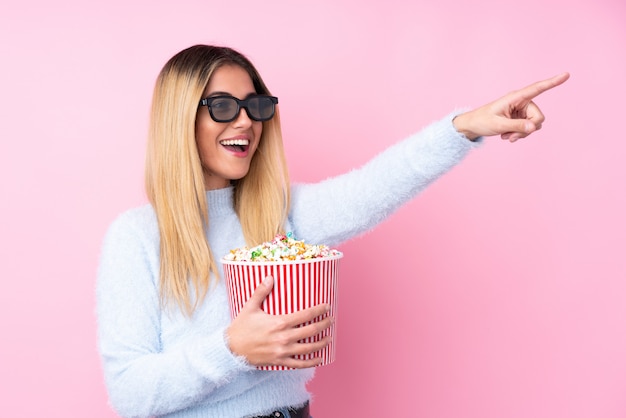 Jeune femme uruguayenne sur mur rose isolé avec des lunettes 3d et tenant un grand seau de pop-corn tout en pointant loin