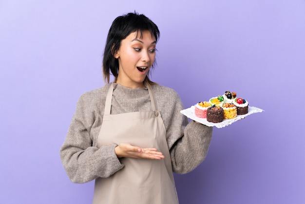 Jeune femme uruguayenne tenant beaucoup de mini gâteaux différents sur violet isolé copyspace holding imaginaire sur la paume