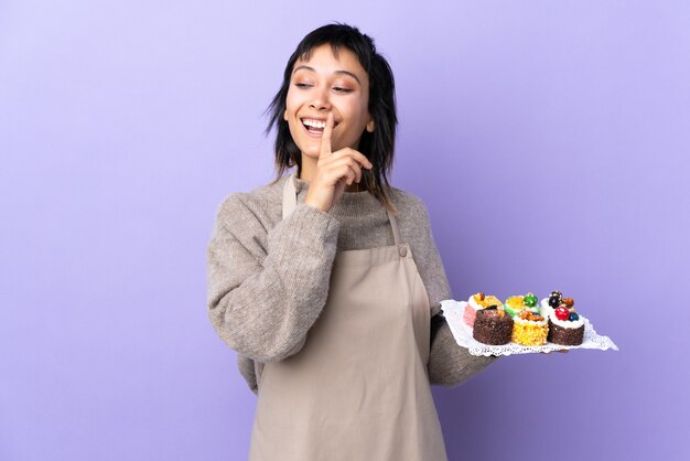 Jeune femme uruguayenne tenant beaucoup de mini gâteaux différents sur violet isolé faisant le geste de silence