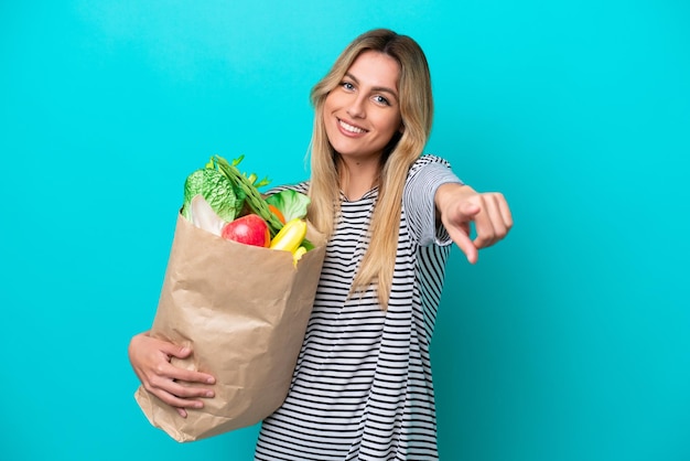 Jeune femme uruguayenne tenant un sac d'épicerie isolé sur fond bleu pointant vers l'avant avec une expression heureuse